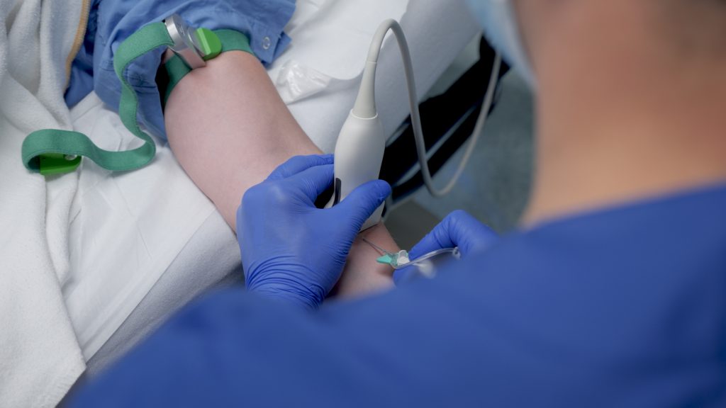 Zorgverlener met blauwe handschoenen prikt in arm van liggende patiënt met stuwband, gebruikte echo voor moeilijke prikkers.
