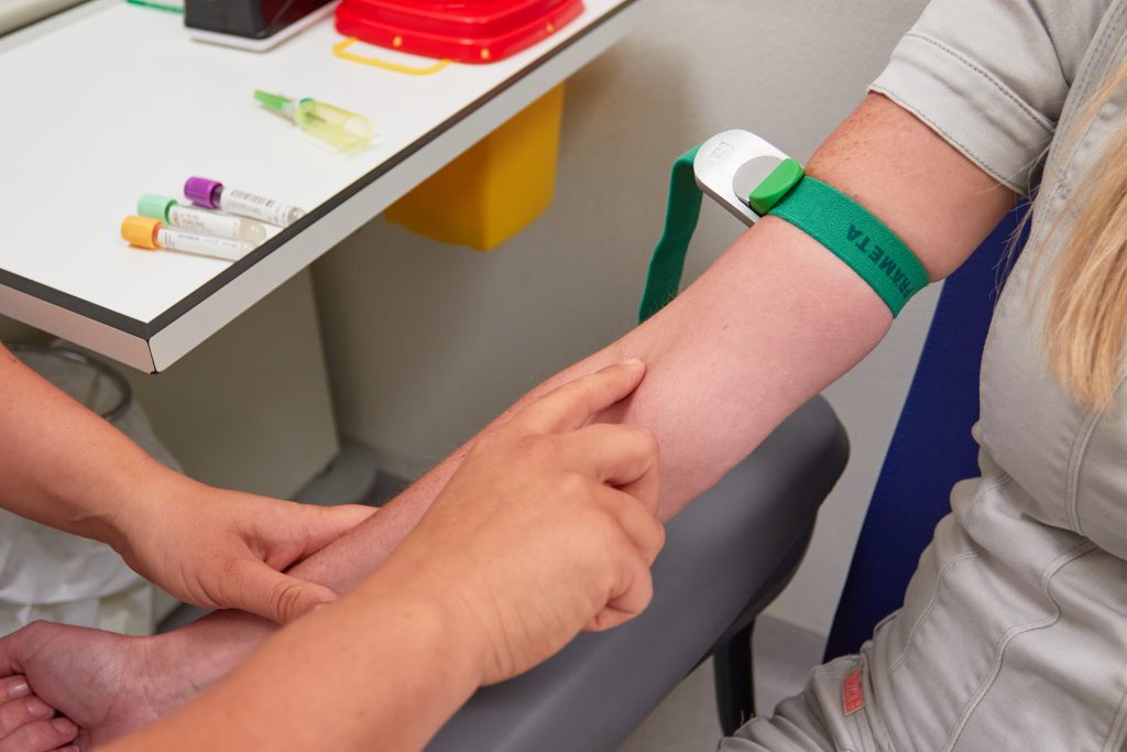 Een zorgmedewerker legt een groene stuwband aan voor bloedafname. Op tafel liggen buisjes en medische benodigdheden.