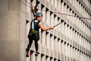 Een persoon in veiligheidsuitrusting ziplinet voor het Catharina Ziekenhuis, met selfie-stick; kabels en harnas zichtbaar.