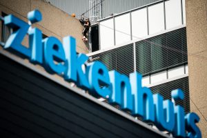 Persoon met veiligheidsuitrusting klimt op ladder aan buitenkant van modern gebouw; vaag blauw bord "Catharina Ziekenhuis" zichtbaar.