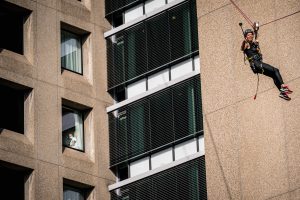 Persoon met helm en harnas op zipline langs hoge kantoorgebouwen; ander kijkt uit raam, Catharina Ziekenhuis op achtergrond.