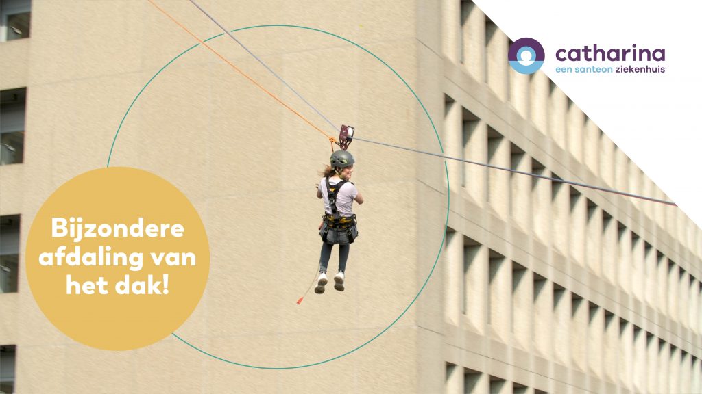 Persoon in veiligheidskleding daalt met een zipline van het dak van een beige gebouw. Catharina Ziekenhuis-logo rechtsboven.