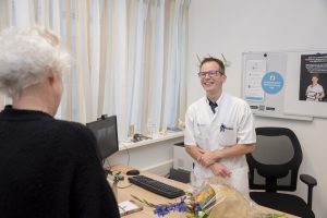Zorgverlener in witte jas praat glimlachend met oudere persoon aan bureau. Computer, bloemen en posters zichtbaar in de kamer.