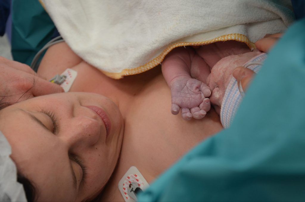 Een pasgeboren baby ligt op de borst van een moeder in het ziekenhuis, onder een wit handdoek. Moeder heeft gesloten ogen.