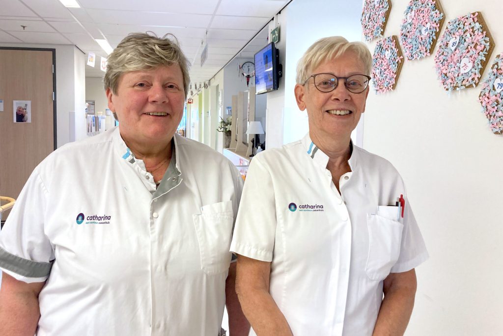 Twee zorgmedewerkers in witte uniformen staan naast elkaar in een lichte bende, glimlachend. Achter hen hangt kunst aan de muur.