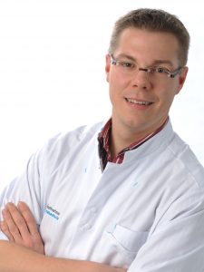 Man met bril en witte labjas staat met gekruiste armen en glimlacht in de camera tegen een witte achtergrond.