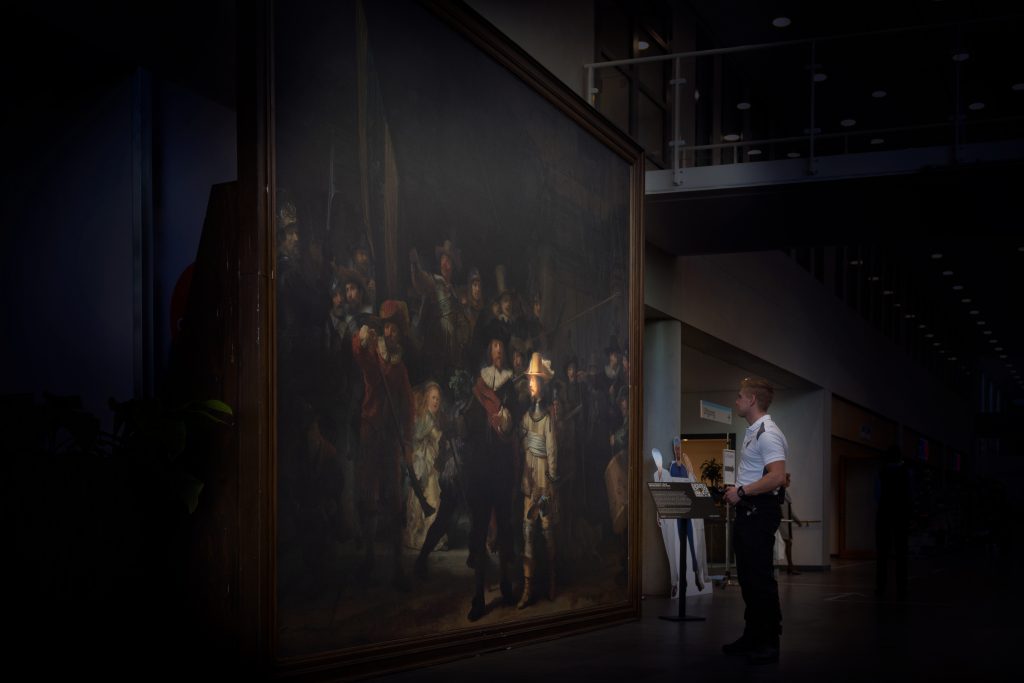 Beveiliger staat voor groot ingelijst Rembrandt-schilderij onder zacht licht in Catharina Ziekenhuis, moderne achtergrond.