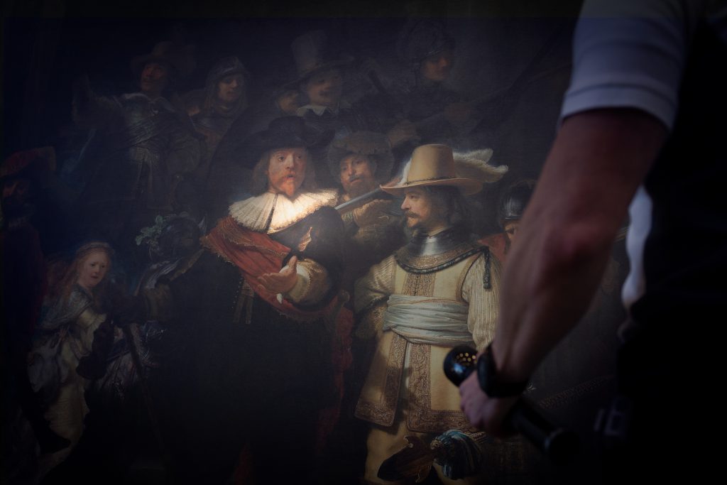 Een persoon met een zaklamp staat voor Rembrandts De Nachtwacht, dat figuren in historische kleding toont.