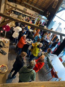 Groep mensen praat bij tafels met drankjes en snacks in een sfeervolle zaal met houten balken. Enkelen dragen gekleurde jassen.