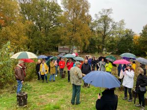 Groep mensen met paraplu's luistert buiten op gras naar spreker in groene jas. Herfstbomen en "meerjema" bord op achtergrond.