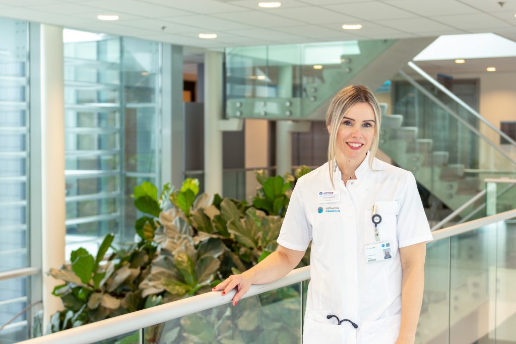 Vrouw in wit uniform met ID-badge, bij planten en reling binnen. Trap en glazen wanden op de achtergrond.