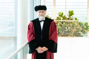 Een oudere man met witte baard en bril draagt een rood-zwarte toga en zwarte baret. Op de achtergrond planten en ramen.