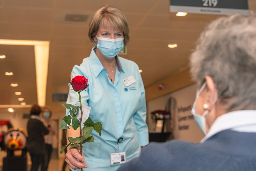 Vrouw in medisch mondkapje en lichtblauw uniform geeft binnenshuis een rode roos aan een andere gemaskerde persoon.