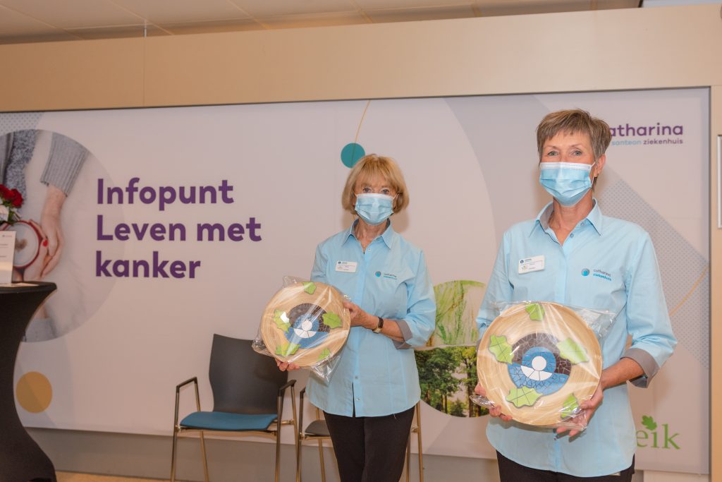 Twee vrouwen met blauwe shirts en mondkapjes houden ronde objecten vast; achter hen een bord: Infopunt Leven met kanker.
