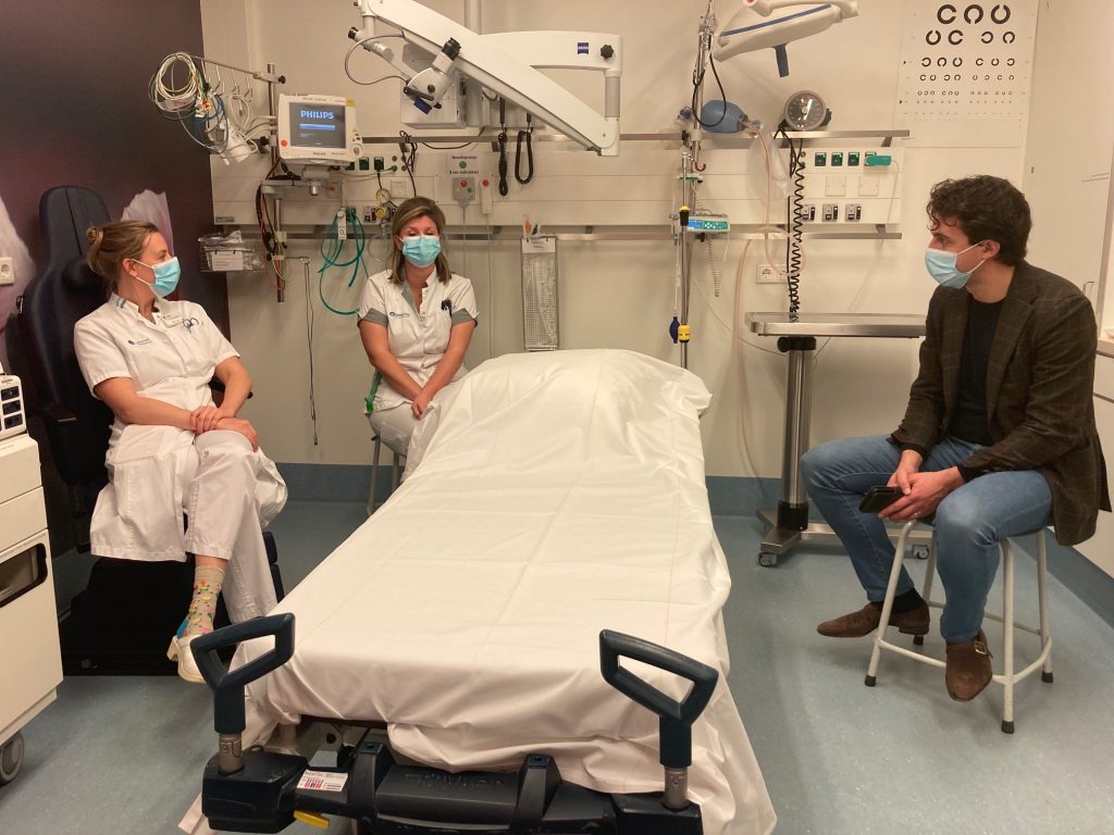 Tijdens een werkbezoek in het Catharina Ziekenhuis zitten drie gemaskerde mensen rond een ziekenhuisbed in een klinische ruimte.