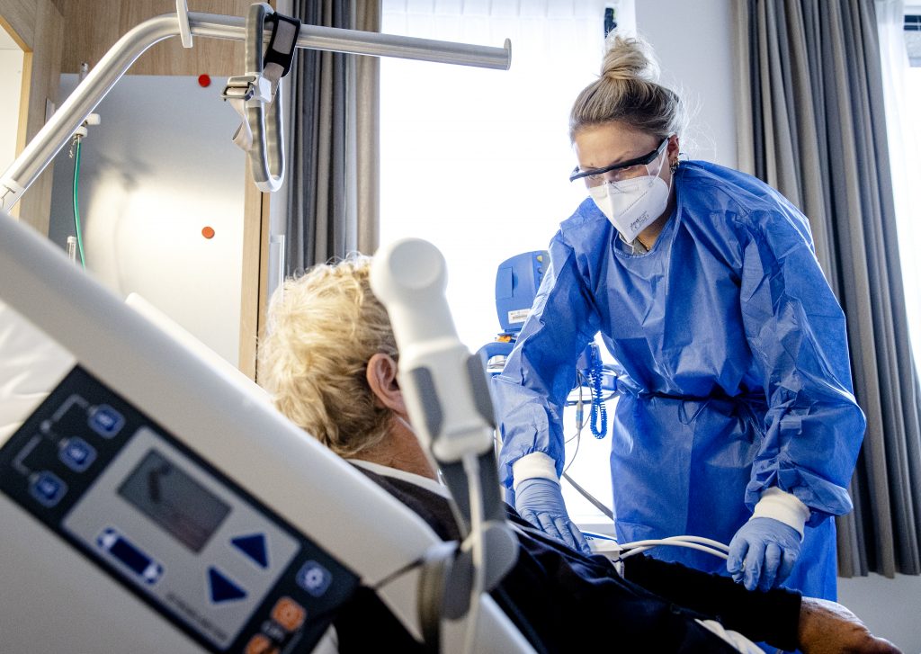 Zorgmedewerker in beschermende kleding controleert patiënt in ziekenhuisbed met medische apparatuur op de achtergrond.