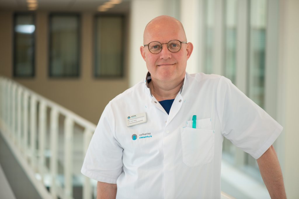 Kale man met bril in wit uniform staat glimlachend in heldere bende, met pennen, IC-dagboek en naamkaartje.