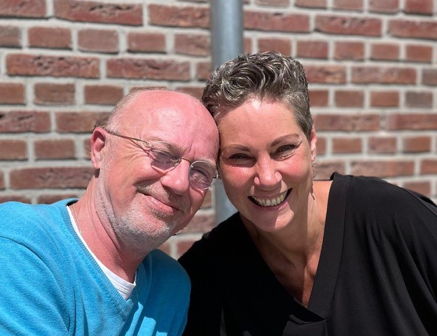 Man met bril in blauw shirt en vrouw in zwart topje zitten naast elkaar buiten voor een bakstenen muur en lachen naar de camera.
