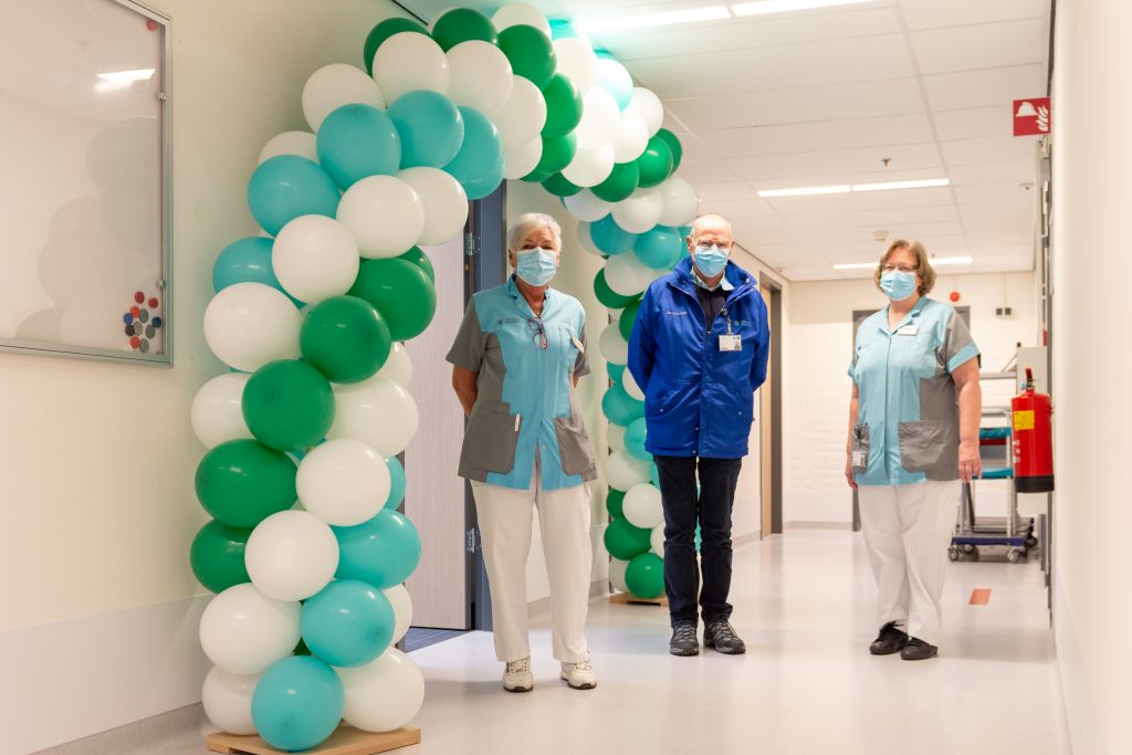 Drie vrijwilligers met mondkapjes en uniform staan in een bende onder groene, witte en blauwe ballonnen, met medische apparatuur.