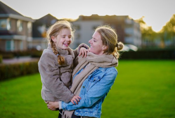 Volwassene houdt lachend kind Fleur vast op grasveld buiten, beiden in jas en sjaal. Huizen en bomen op achtergrond.