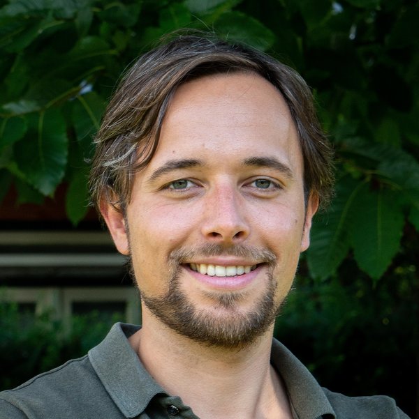 Man met kort bruin haar en baard in donkergroen overhemd glimlacht naar de camera; vage groene planten en raam op achtergrond.