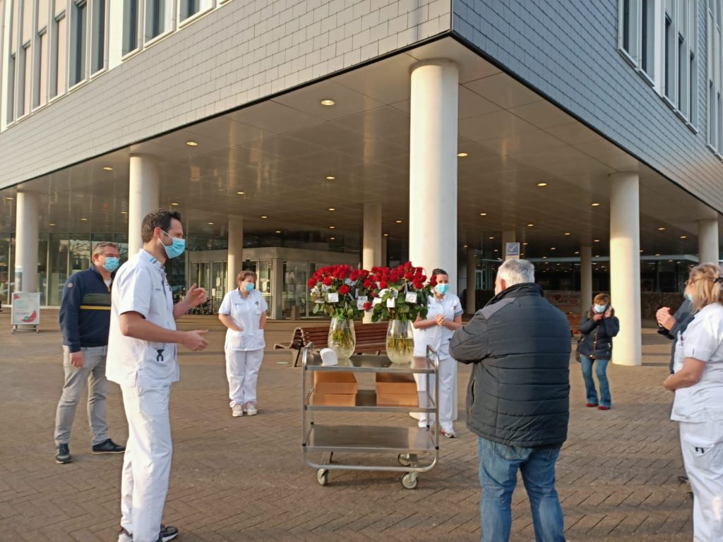 Een groep mensen, waaronder zorghelden in witte uniformen en mondkapjes, staat bij een kar met rozen voor een modern gebouw.
