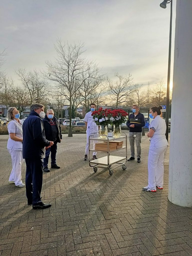Zorghelden, deels in witte uniformen en mondkapjes, staan buiten bij een kar met rozen. Bomen zonder blad en auto's op de achtergrond.