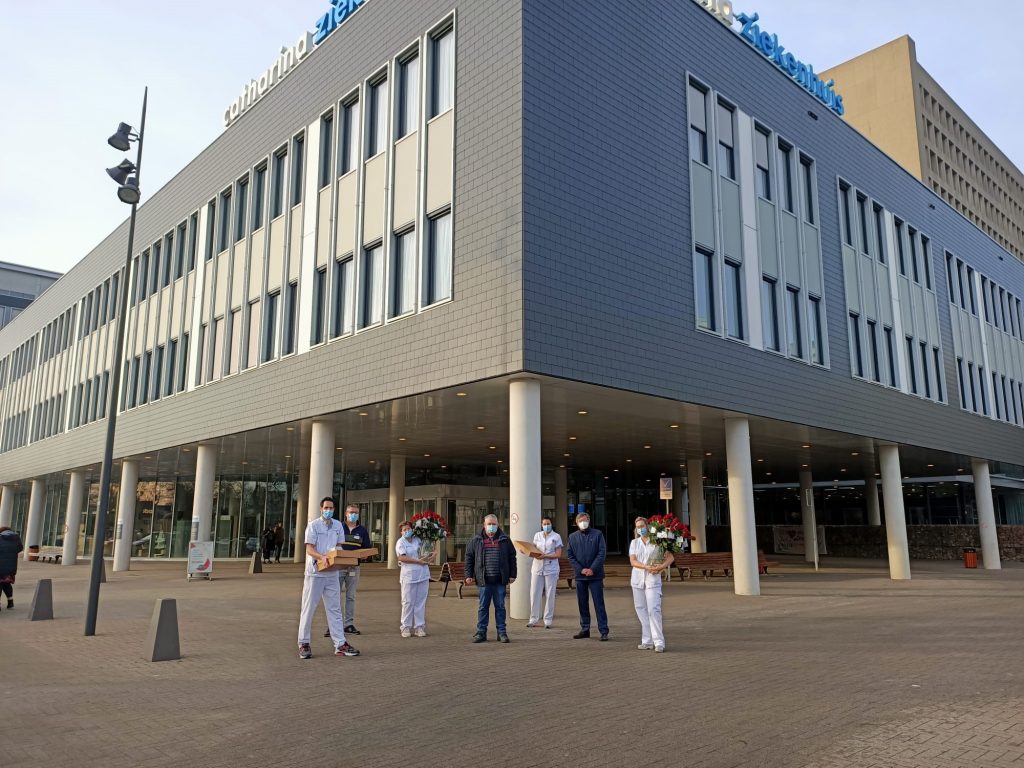 Zes personen, waaronder drie zorghelden in witte uniformen met bloemen, staan buiten bij een modern gebouw met grote ramen.