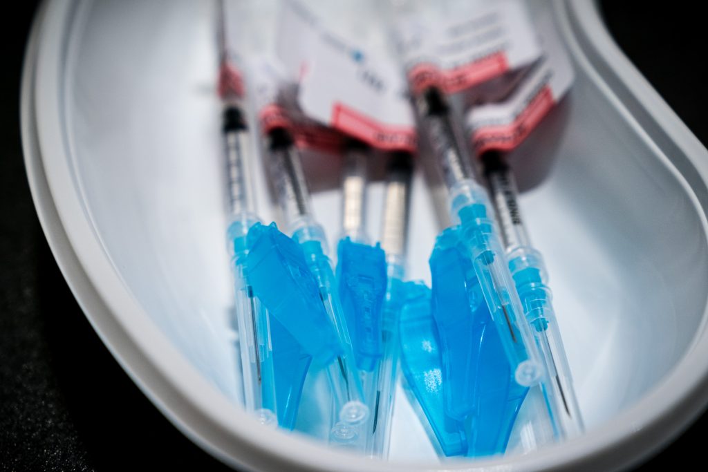 Vier spuiten met blauwe dop in een witte nierbekken, klaar voor extra vaccinatie van kwetsbare patiënten, met rood-witte etiketten.