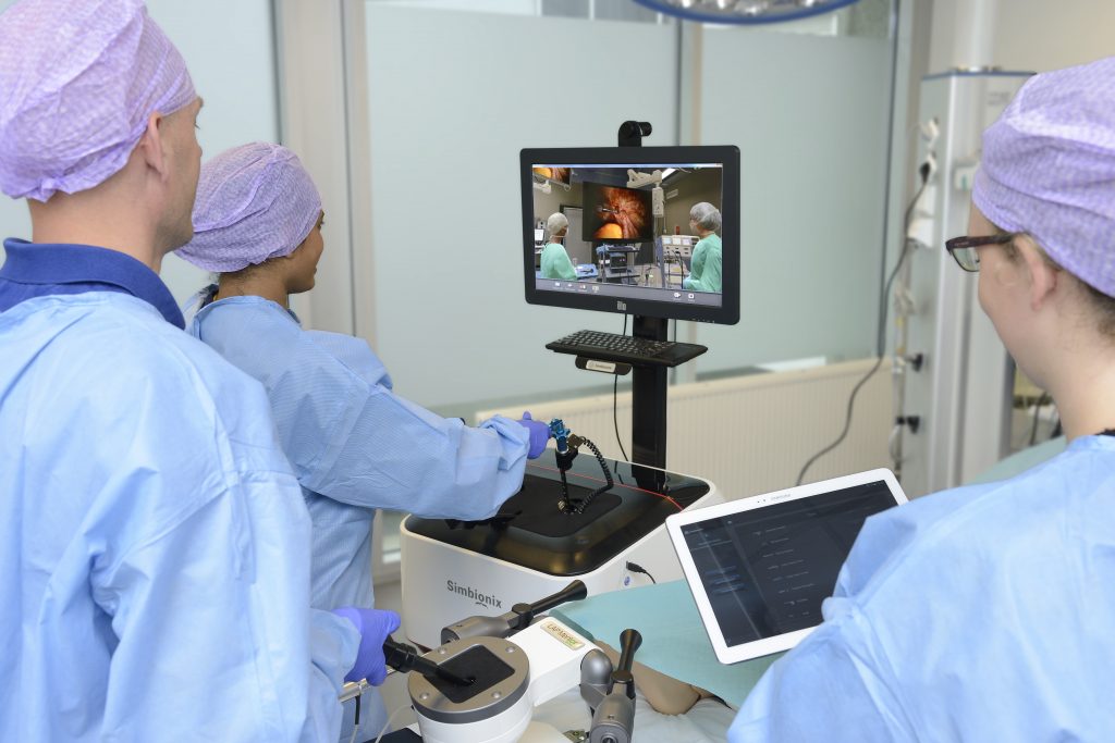 Drie medische professionals in operatiekleding werken met een operatiesimulator; één bedient, één observeert, en één houdt een tablet vast.
