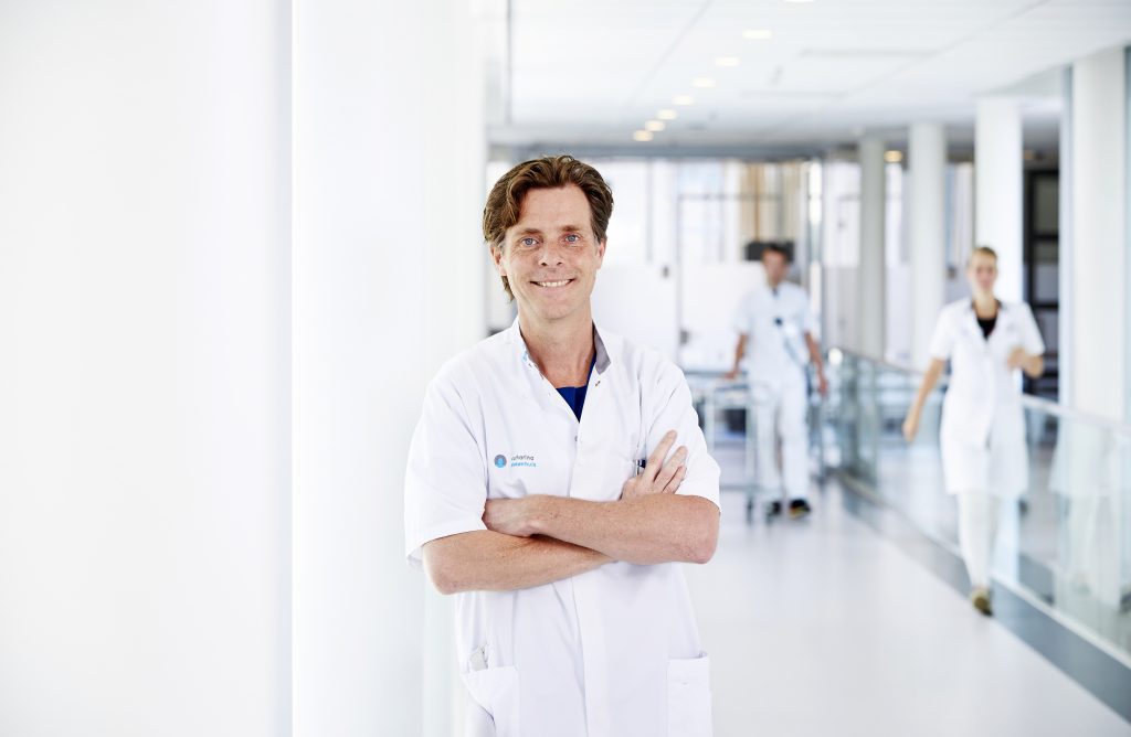 Zorgprofessional in witte jas staat met gekruiste armen in heldere gang; twee collega's lopen op de achtergrond.