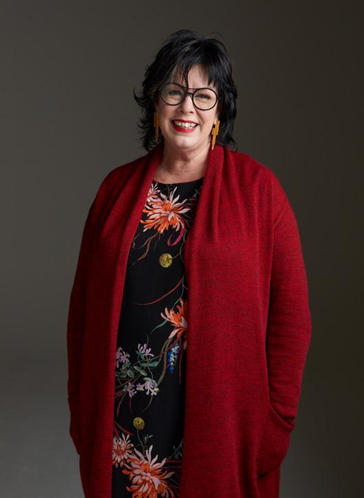 Lizette, met kort donker haar en bril, glimlacht. Ze draagt een rode vest, zwarte jurk met bloemen en oranje oorbellen.
