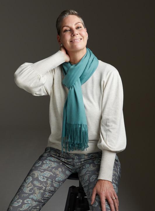 Renate Spijkerman met uitgezaaide eierstokkanker zit op een kruk in witte trui, gekleurde broek en blauwe sjaal, hand aan haar nek.
