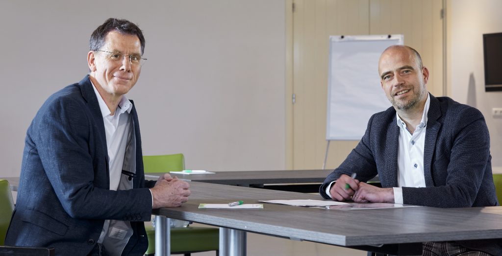 Twee mannen in pak aan tafel in een conferentieruimte in Weert, met papieren en pennen. Flip-over en groene stoelen op achtergrond.