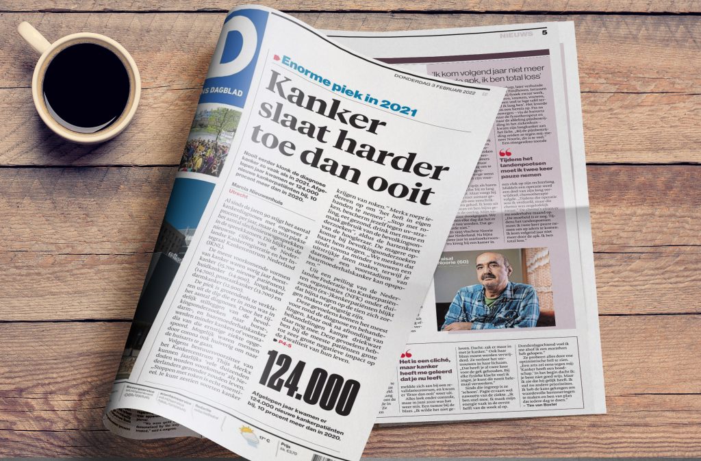 Nederlandse krant open op houten tafel naast kop koffie. Kop: "Kanker slaat harder toe dan ooit." Foto man in ruitoverhemd.