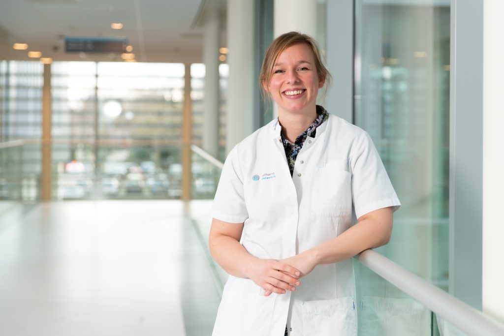 Vrouw in witte jas leunt glimlachend tegen glazen reling in lichte bende met grote ramen, symbool voor Nederlandse fysiotherapie.