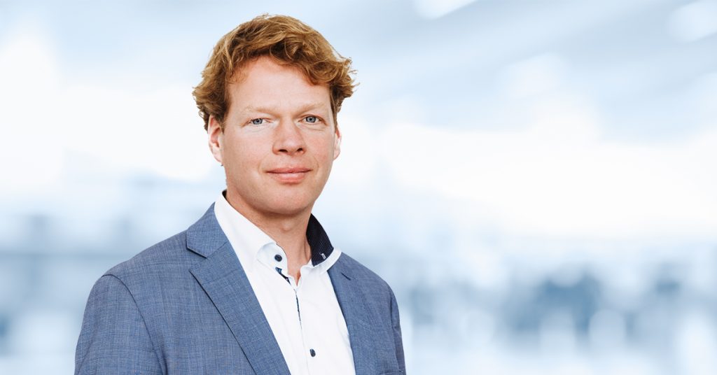 Man met kort, goudkleurig rossig-blond haar in lichtblauw pak en wit shirt staat neutraal kijkend voor een vage, lichte achtergrond.