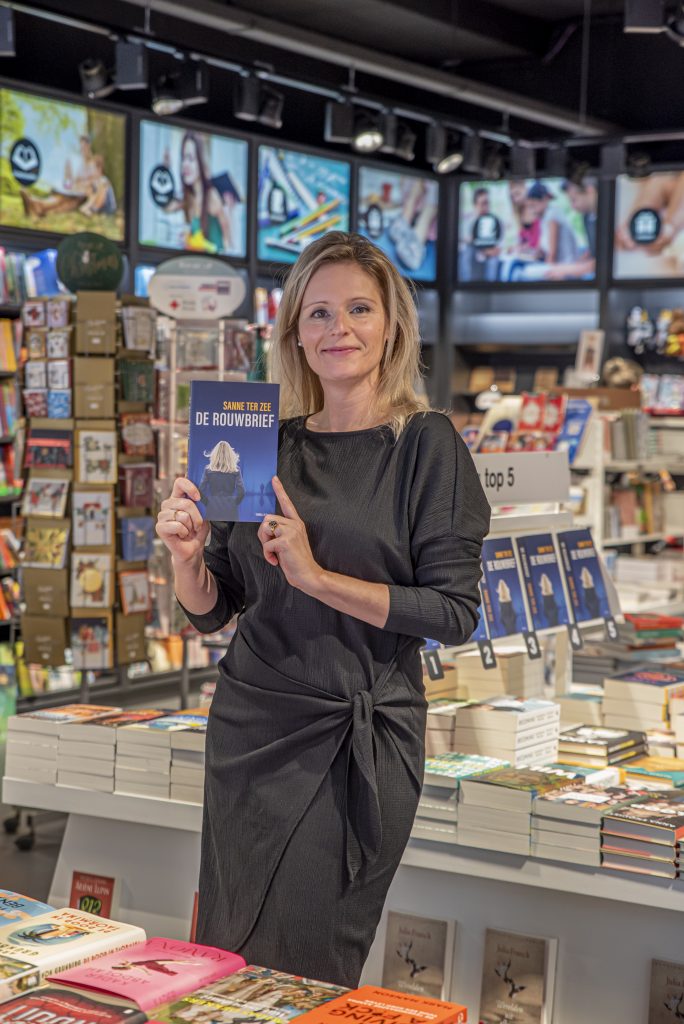 Een vrouw in een zwarte jurk houdt het boek "De Rouwbrief" vast in een boekwinkel met stapels boeken en volle boekenkasten.