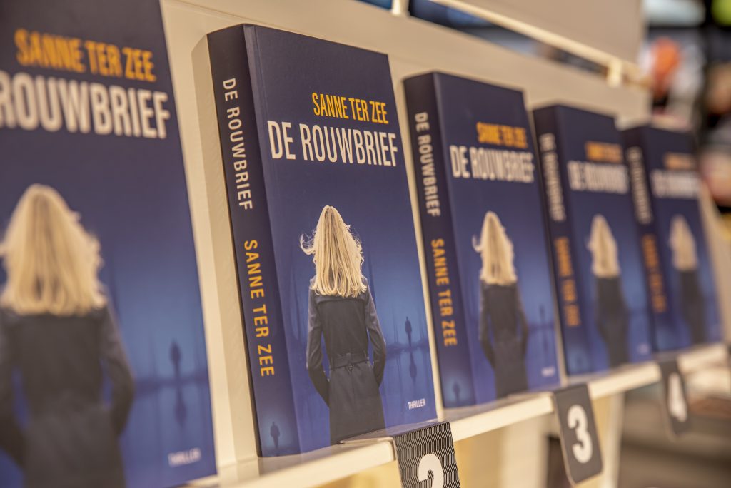 Meerdere exemplaren van De Rouwbrief van Sanne Ter Zee op een plank, met op de omslag een blonde vrouw gezien van achteren.