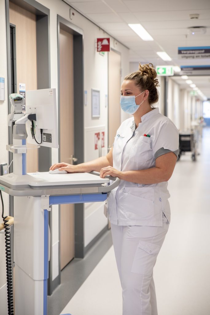 Een verpleegkundige met mondmasker en wit uniform staat bij een computer in een lichte ziekenhuisgang met medische apparatuur.