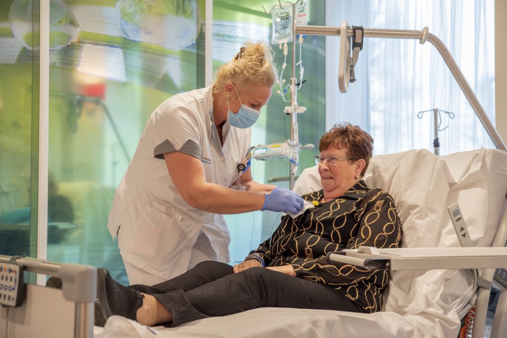 Een verpleegkundige met mondmasker en handschoenen helpt een oudere vrouw op een ziekenhuisbed, met medische apparatuur op de achtergrond.
