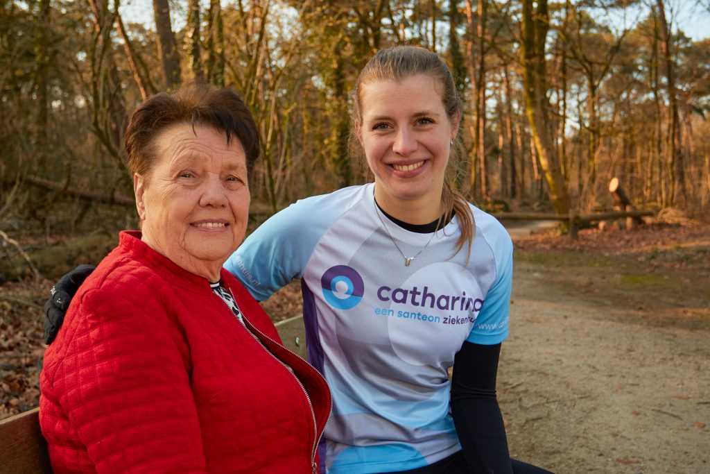 Een oudere vrouw in rood jack zit op een bank naast Suzanne in Catharina ziekenhuisshirt. Ze zijn buiten en lachen.
