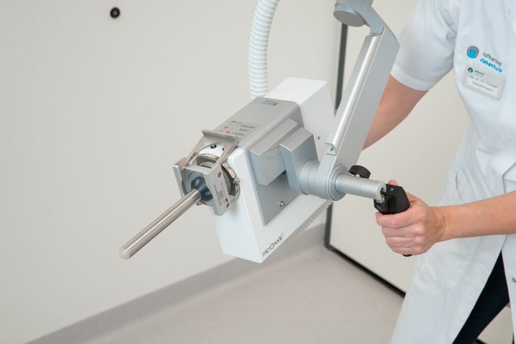 Een persoon in een witte jas bedient een medisch beeldvormingsapparaat met metalen staaf en slang in een klinische omgeving.