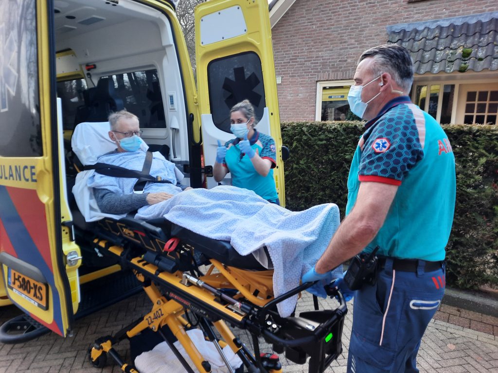 Oudere met mondmasker op brancard wordt door twee ambulancemedewerkers naar ambulance bij bakstenen gebouw gebracht.