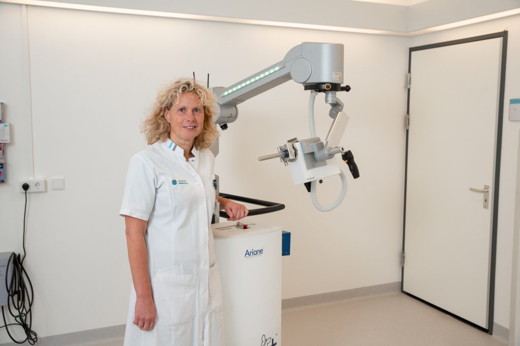 Persoon in wit uniform naast groot apparaat voor behandeling van endeldarmkanker in witte kliniek, deur op achtergrond.