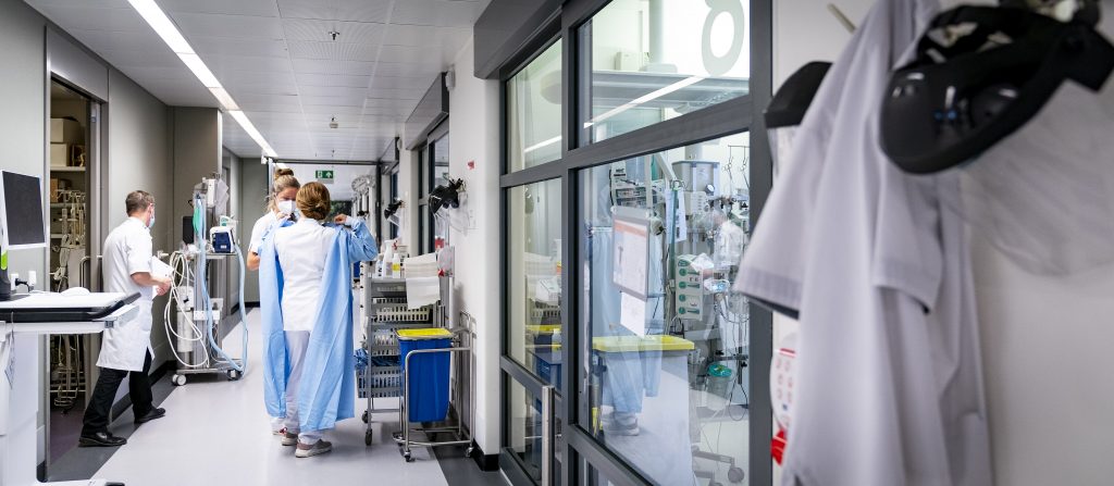 Ziekenhuisgang met medisch personeel in beschermende kleding, patiënten en apparatuur; jasjes hangen aan de muur.
