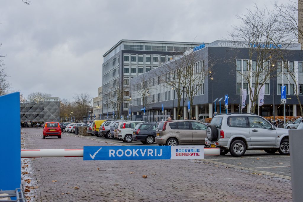 Parkeerplaats met auto's, witte rookvrije-barrière met blauwe borden. Kantoorgebouwen op de achtergrond, bewolkte dag.