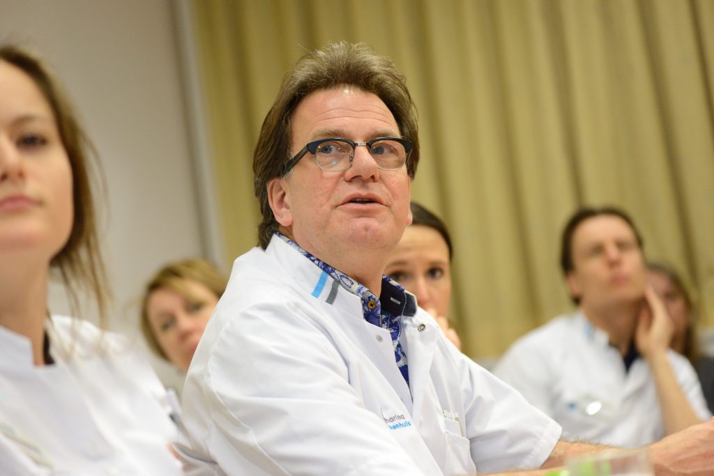 Een man met bril en witte jas zit in een vergaderruimte met collega's; anderen zijn deels onscherp zichtbaar.