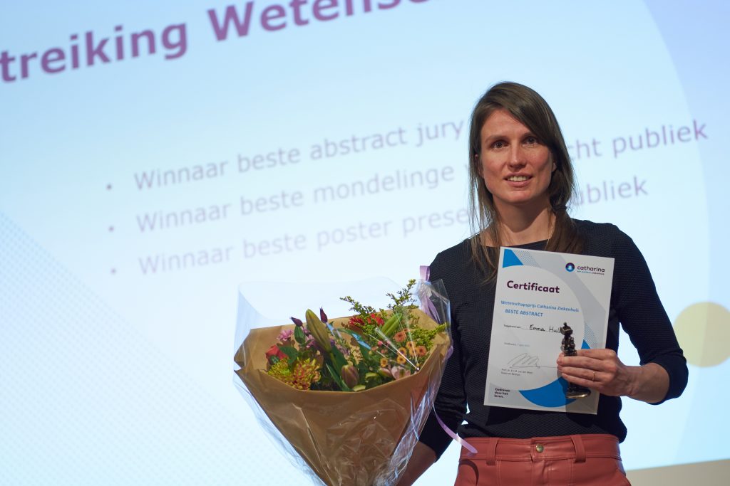 Vrouw met bloemen en certificaat voor scherm met prijsinformatie tijdens Catharina Wetenschapsavond, erkenning Emma Hulshof.