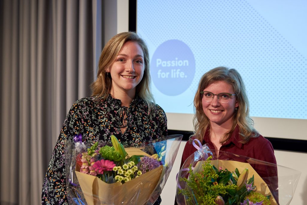 Twee mensen met bloemen poseren en lachen. Op de achtergrond een scherm met "Passion for life." bij Catharina Wetenschapsavond 2022.